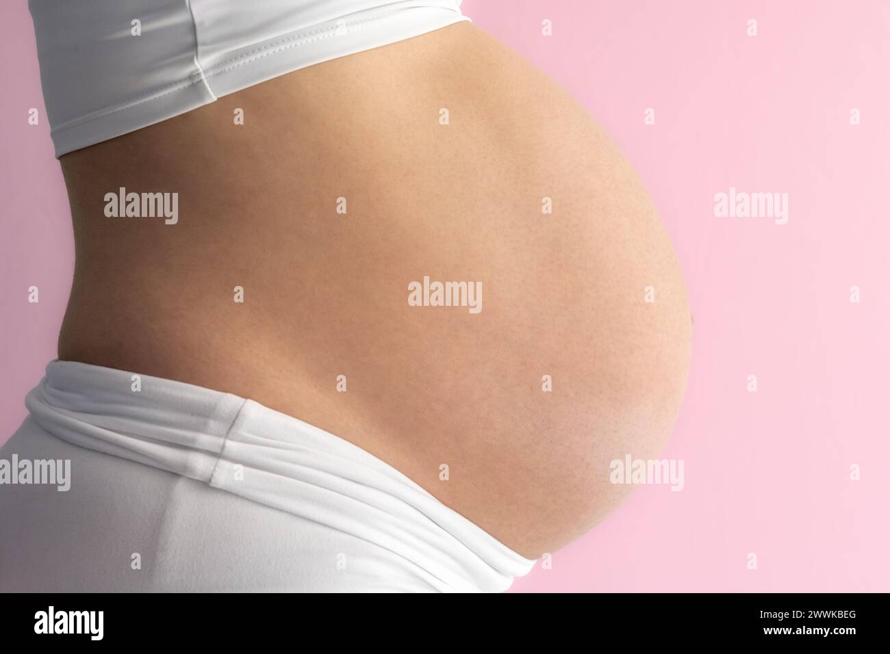 Description: Midsection of unrecognizable standing mother with very round pregnant baby belly. Side view. Pink background. Bright shot. Stock Photo