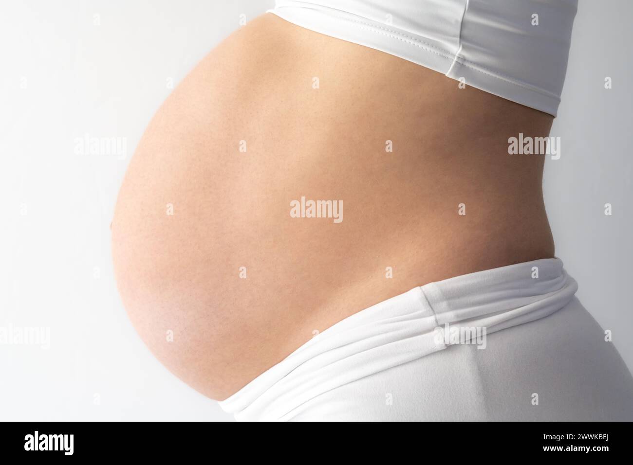Description: Middle part of unrecognizable standing mother with very round pregnant baby bump. Side view. White background. Bright shot. Stock Photo