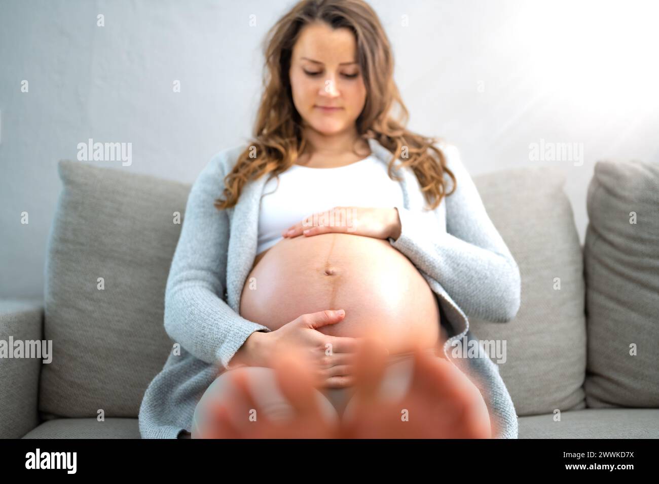 Description: Frontal view of happy woman sitting on sofa gently holding her belly in expectation of having a baby in last stage of pregnancy. Pregnanc Stock Photo
