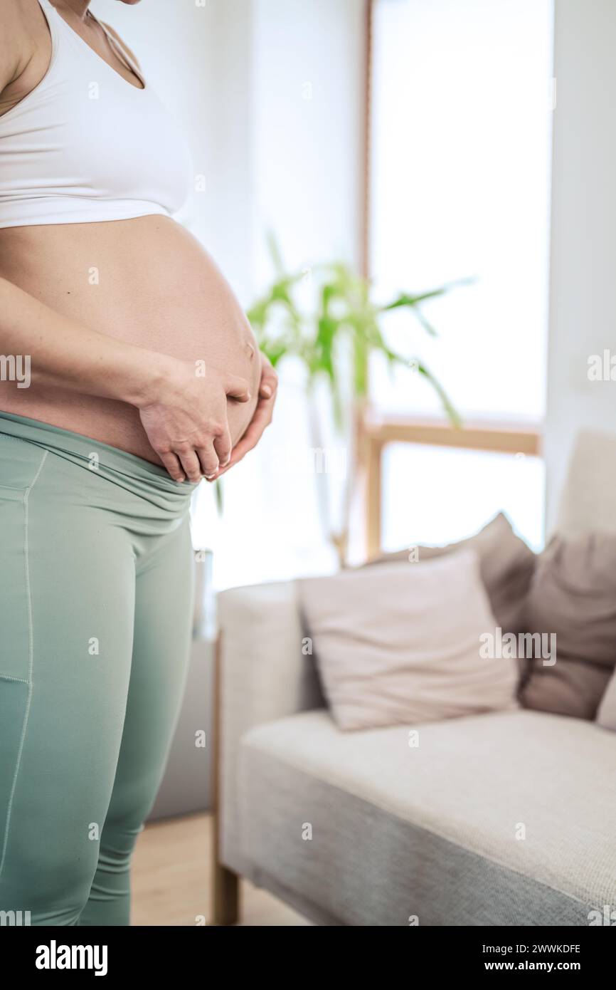 Description: Lateral view of unrecognizable woman gently holding her belly in final months of pregnancy. Pregnancy first trimester - week 18. Side vie Stock Photo