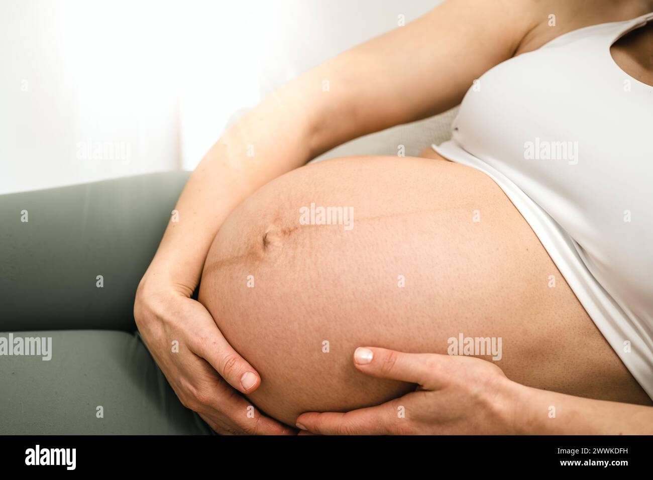 Description: View of woman lying on her side on a sofa gently holding her belly in the last stage of pregnancy. Pregnancy third trimester - week 34. S Stock Photo