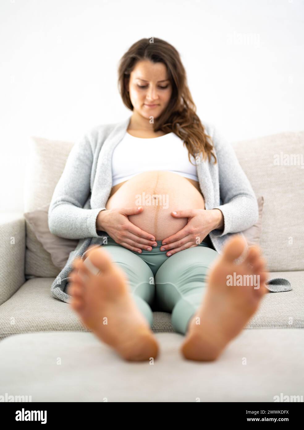 Description: Frontal view of smiling woman sitting on a sofa proudly holding her belly in expectation of the baby during the last stage of pregnancy. Stock Photo