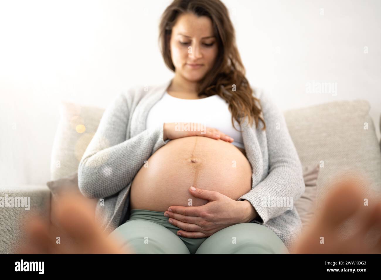 Description: Frontal view of woman sitting on a sofa gently holding her belly in expectation of the baby during the last stage of pregnancy. Pregnancy Stock Photo