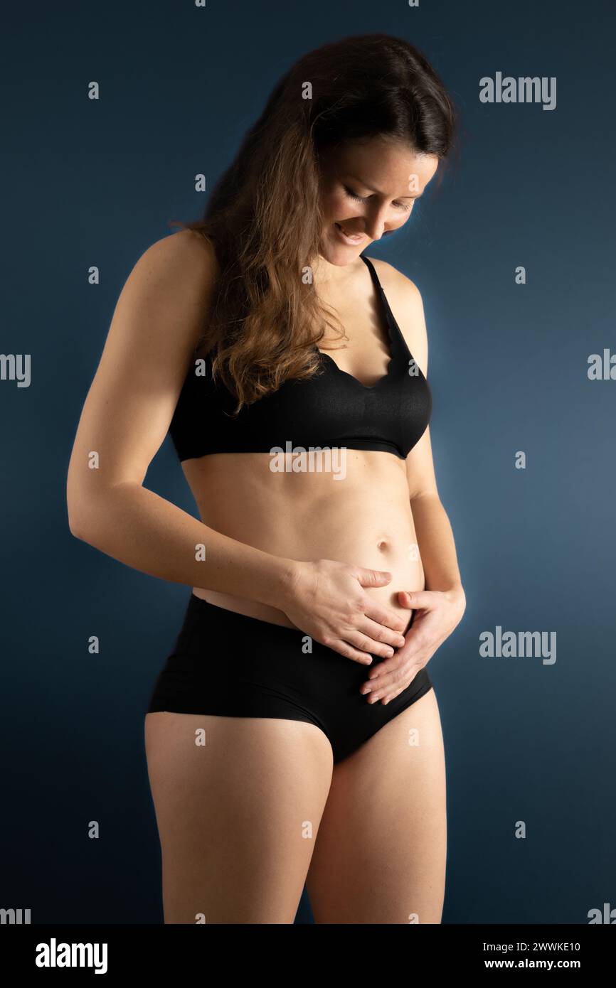 Description: Side angle view of smiling woman holding her belly gently during first months of pregnancy. Pregnancy first trimester - week 18. Side vie Stock Photo