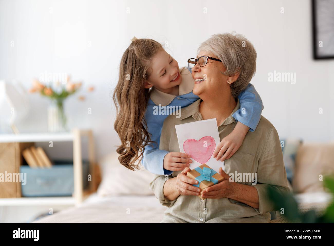 Beautiful cute child congratulating grandmother and giving her postcard. Woman and girl are hugging and laughing. Stock Photo