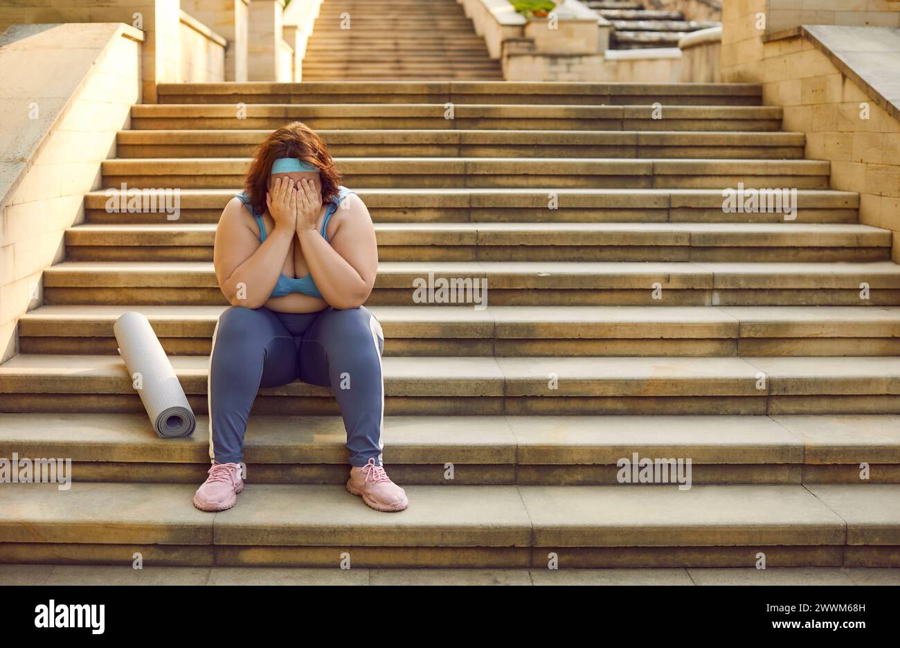 Sad tired young overweight woman with sports mat sitting on stone steps and crying Stock Photo