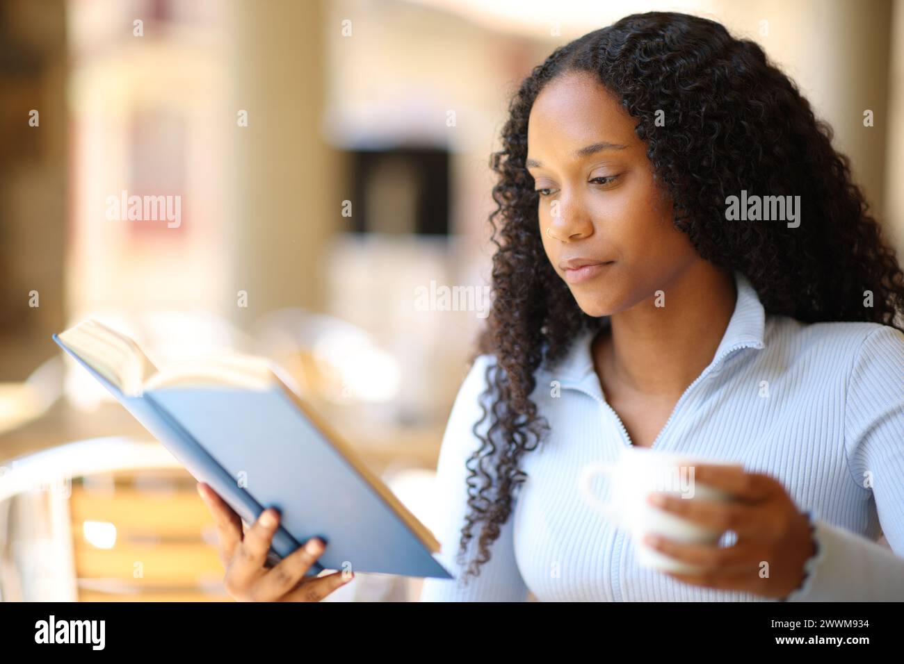 Black woman reading a paper book drinking coffee in a restaurant terrace Stock Photo