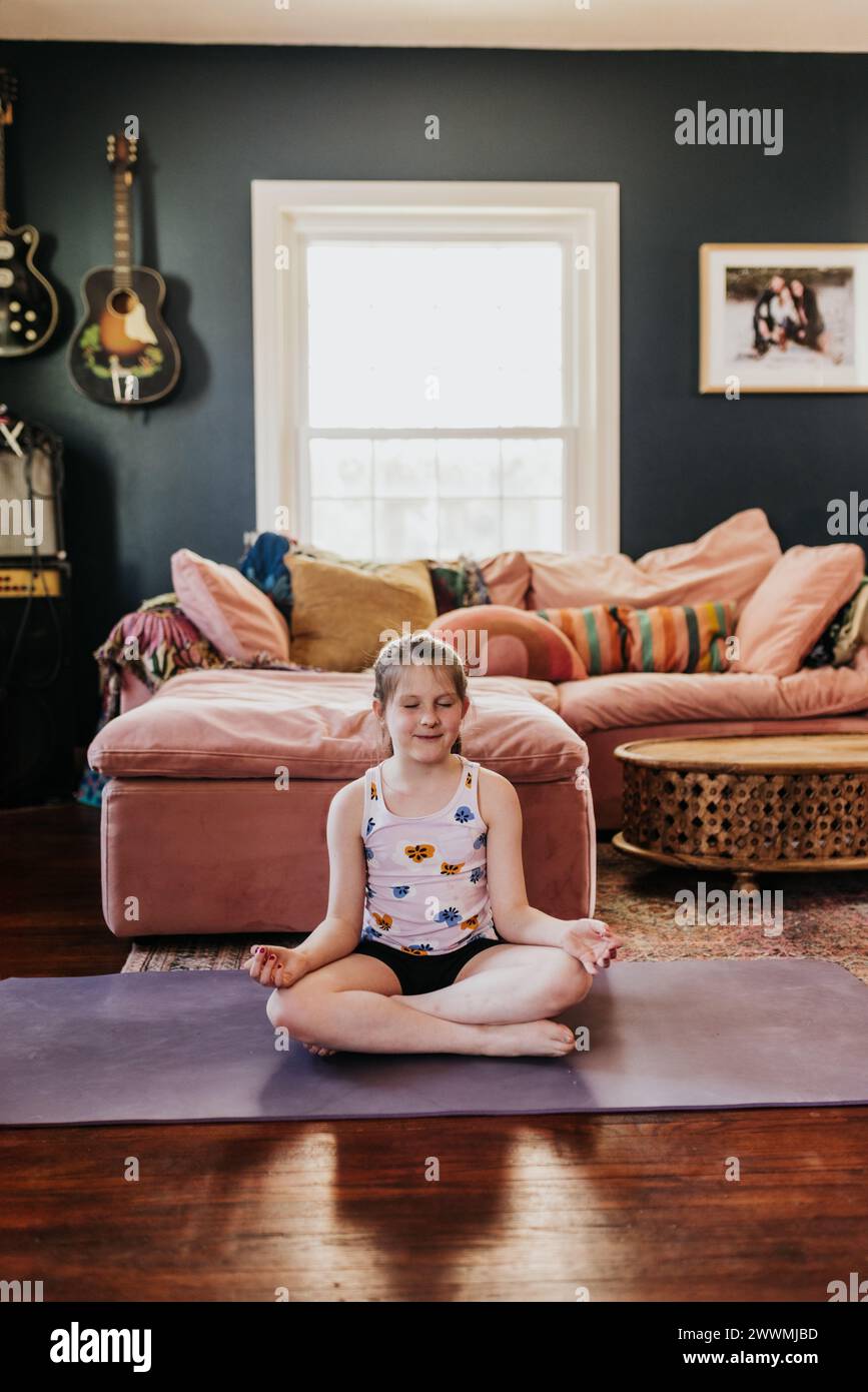 Young girl meditates while sitting on purple yoga mat in living room Stock Photo