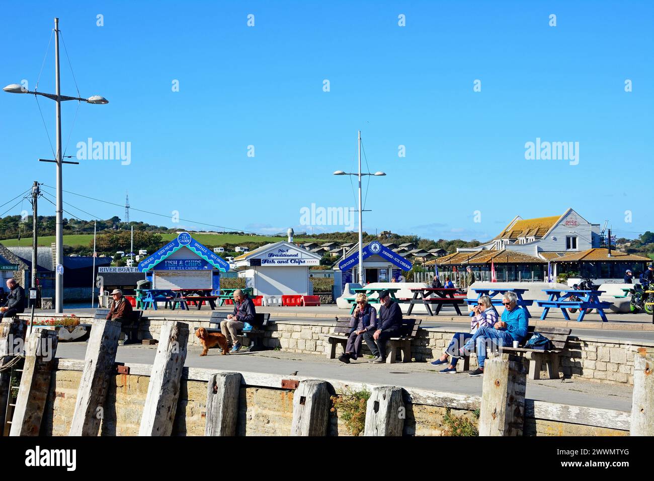 Tourists relaxing on benches in the harbour area with The Rise restaurant and snack kiosks to the rear, West Bay, Dorset, UK. Stock Photo