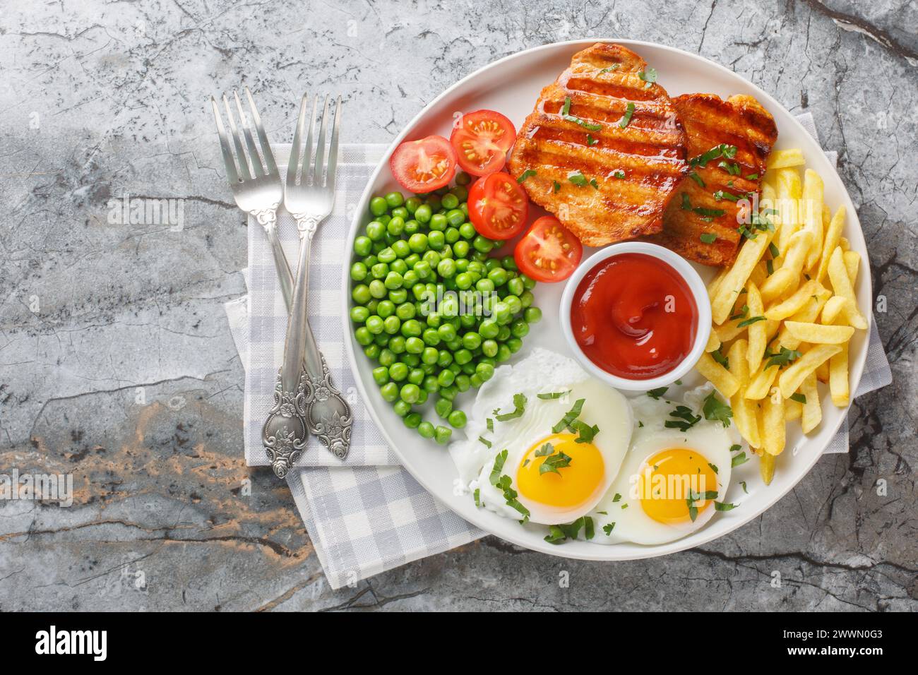 Fried pork loin steak served with French fries, fied eggs, green pea and fresh tomato closeup on the plate on the table. Horizontal top view from abov Stock Photo