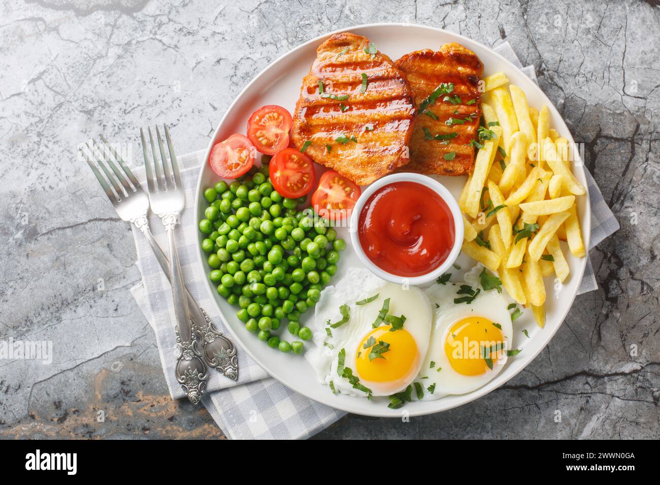 Tender grilled steak served with crisp golden French fries, fied eggs, green pea and fresh tomato closeup on the plate on the table. Horizontal top vi Stock Photo