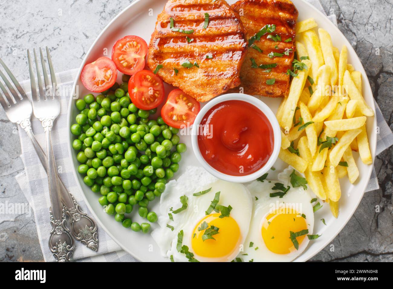 Hearty dinner of grilled loin steak with green peas, fried eggs, french fries, fresh tomatoes and sauce close-up in a plate on the table. Horizontal t Stock Photo
