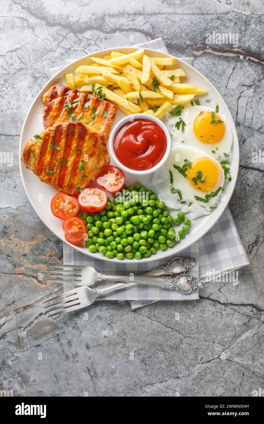 Delicious grilled loin steak with green peas, fried eggs, french fries, tomatoes and sauce close-up in a plate on the table. Vertical top view from ab Stock Photo