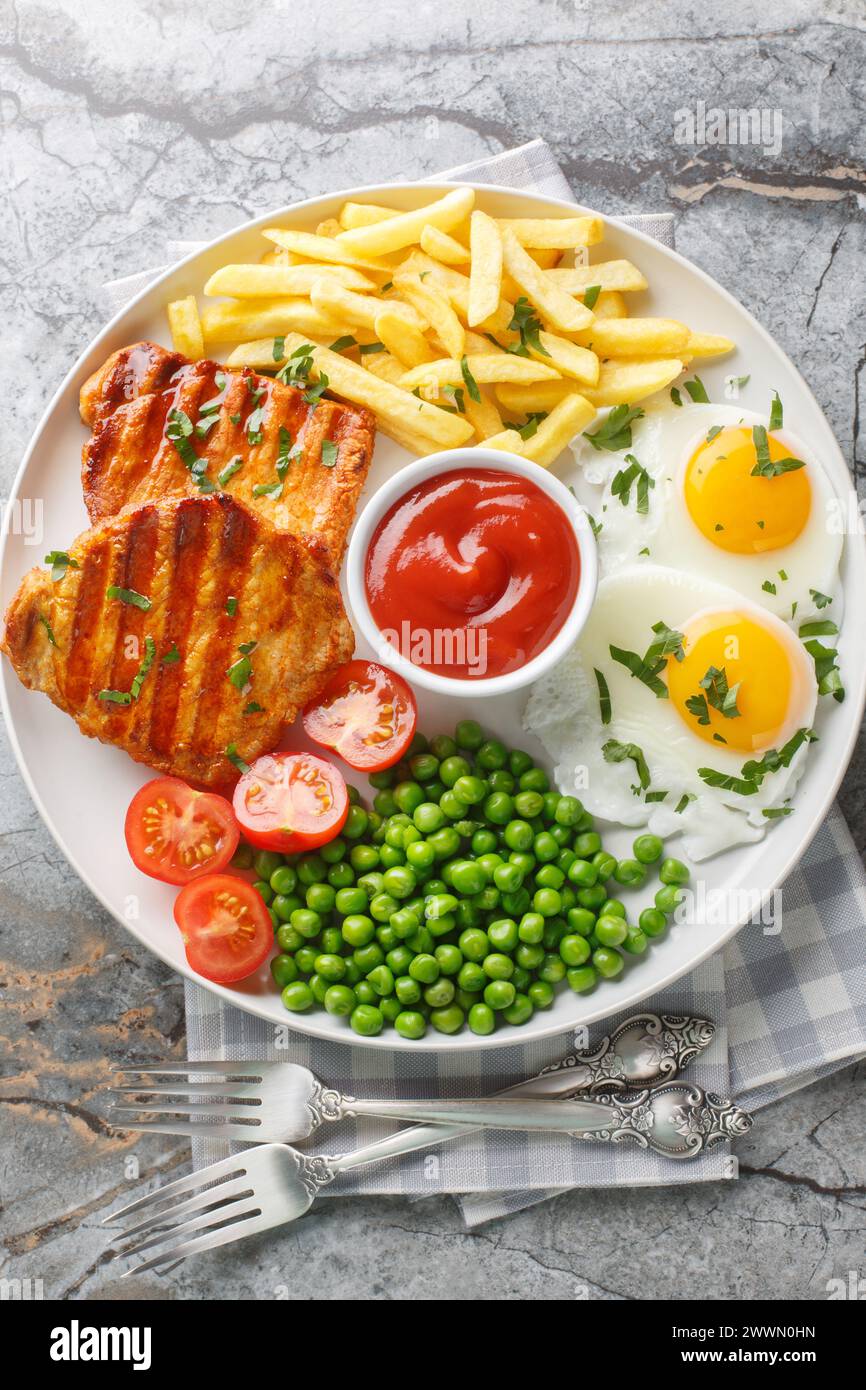Set lunch Grilled steak with green peas, fried egg, french fries, tomatoes and sauce close-up in a plate on the table. Vertical top view from above Stock Photo