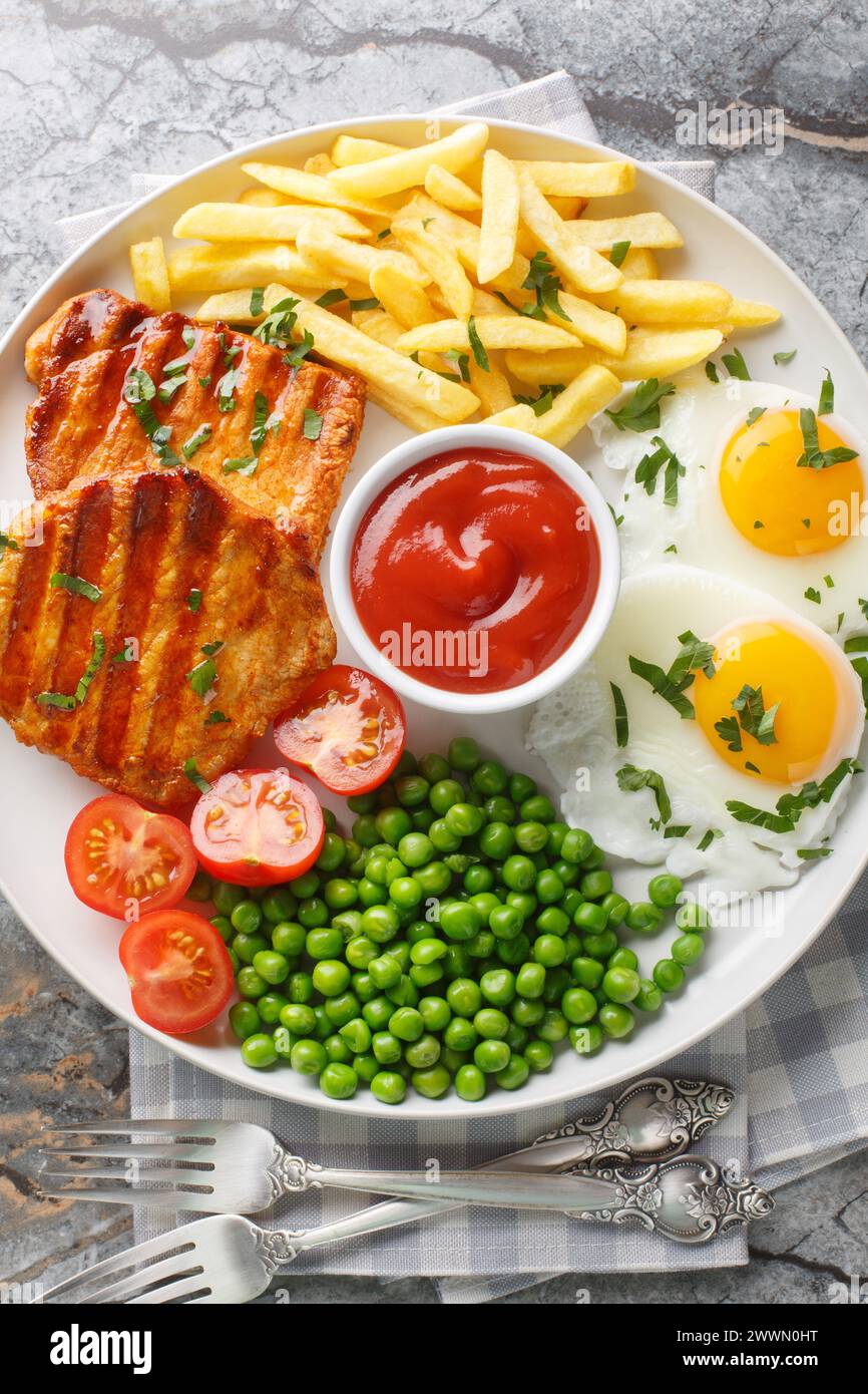 Grilled steak with green peas, fried egg, french fries, tomatoes and sauce close-up in a plate on the table. Vertical top view from above Stock Photo
