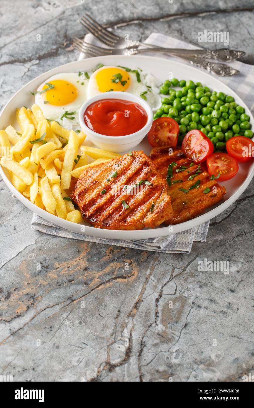 Delicious grilled loin steak with green peas, fried eggs, french fries, tomatoes and sauce close-up in a plate on the table. Vertical Stock Photo