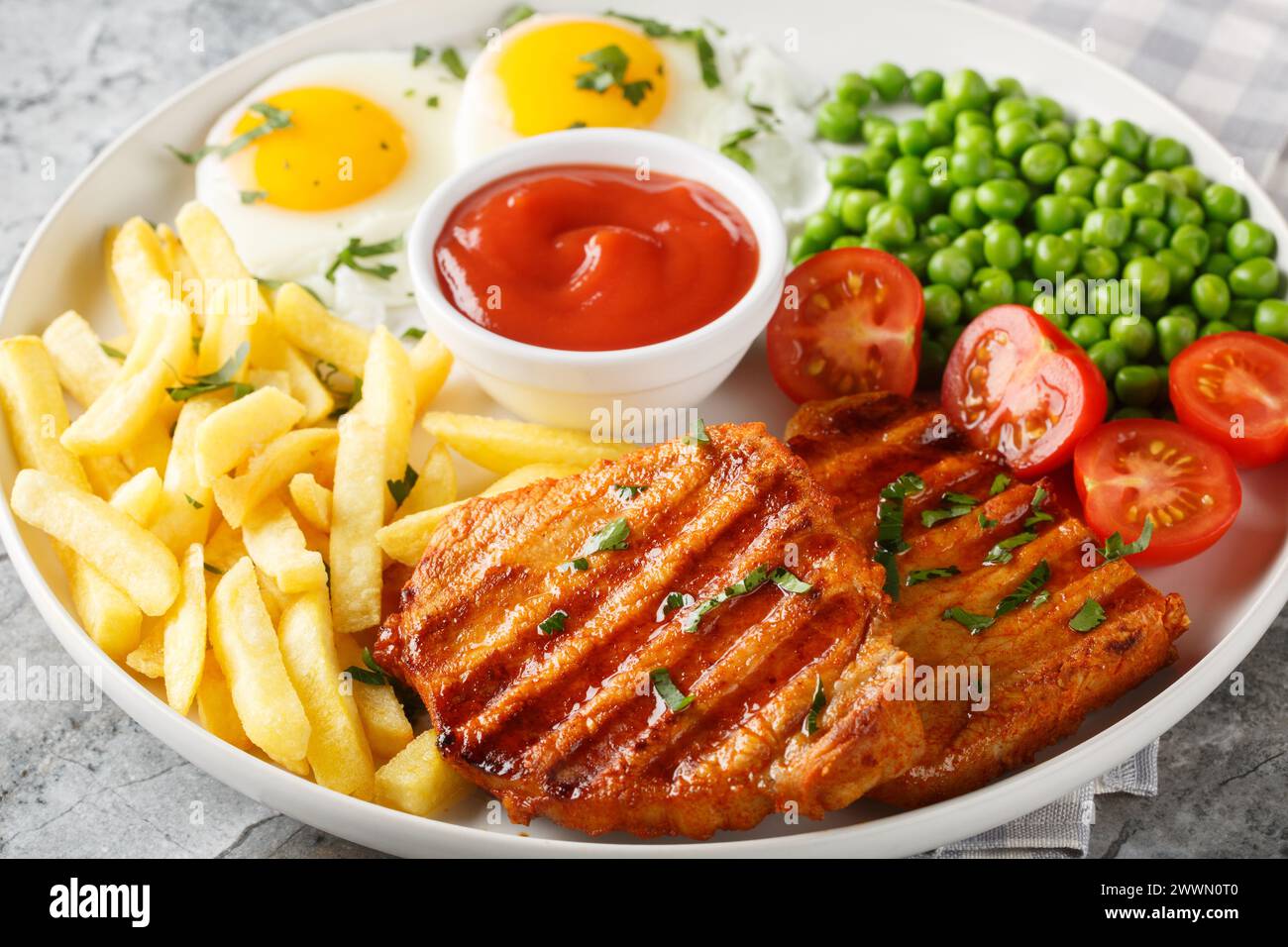 Fried pork loin steak served with French fries, fied eggs, green pea and fresh tomato closeup on the plate on the table. Horizontal Stock Photo