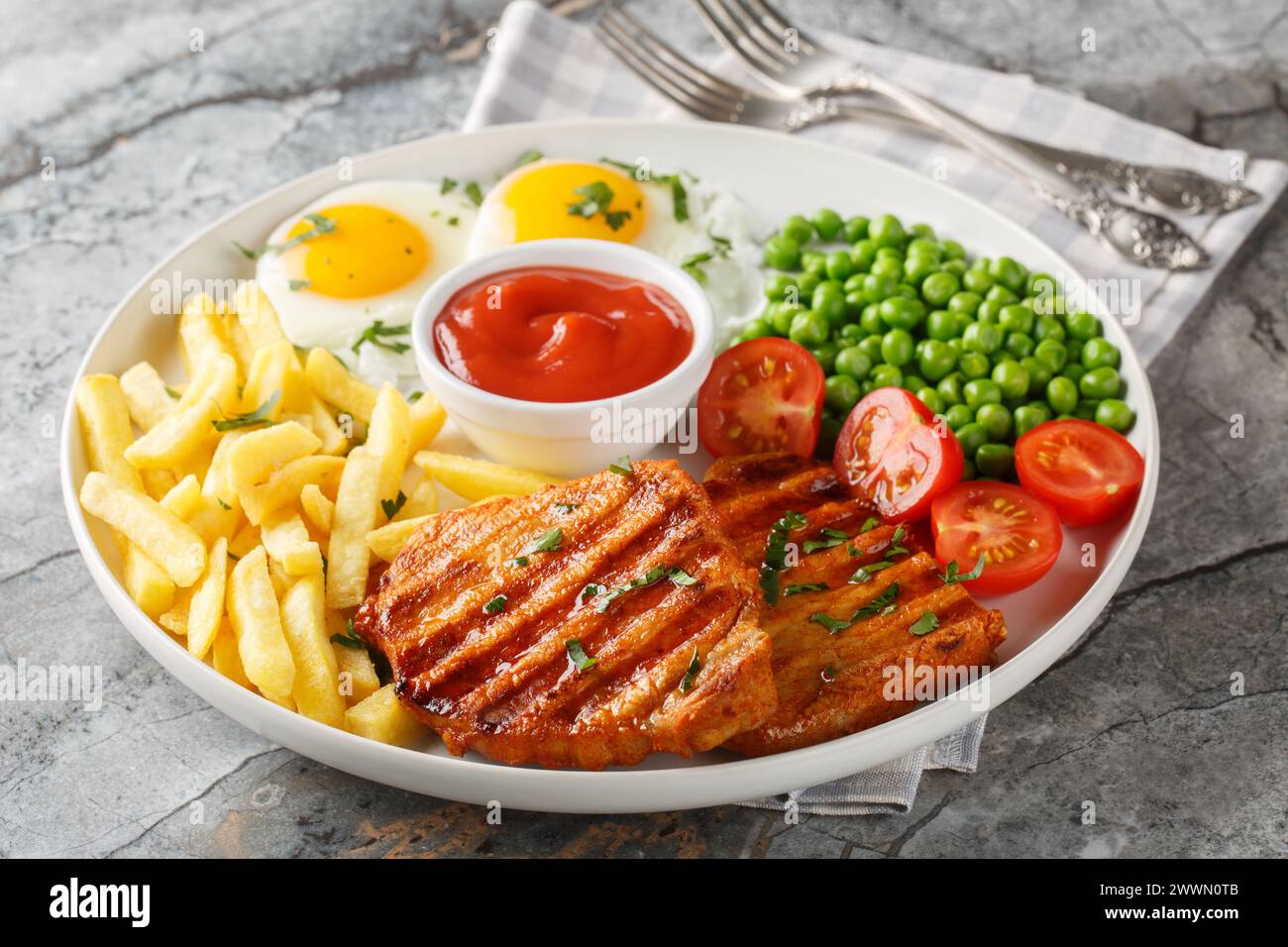 Tender grilled steak served with crisp golden French fries, fied eggs, green pea and fresh tomato closeup on the plate on the table. Horizontal Stock Photo