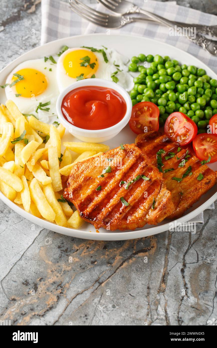 Grilled steak with green peas, fried egg, french fries, tomatoes and sauce close-up in a plate on the table. Vertical Stock Photo