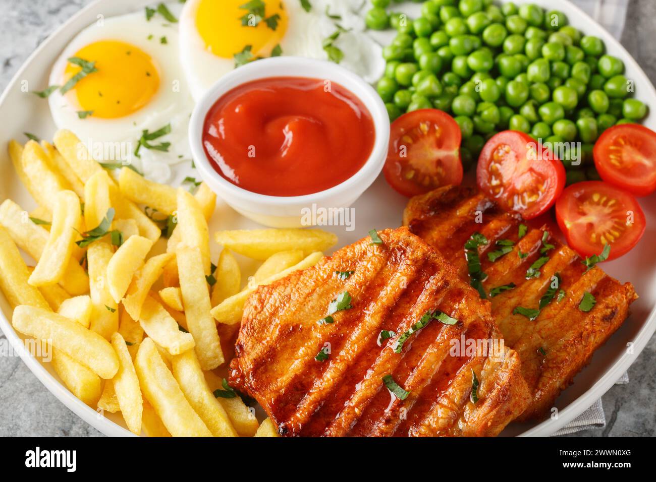 Hearty dinner of grilled loin steak with green peas, fried eggs, french fries, fresh tomatoes and sauce close-up in a plate on the table. Horizontal Stock Photo