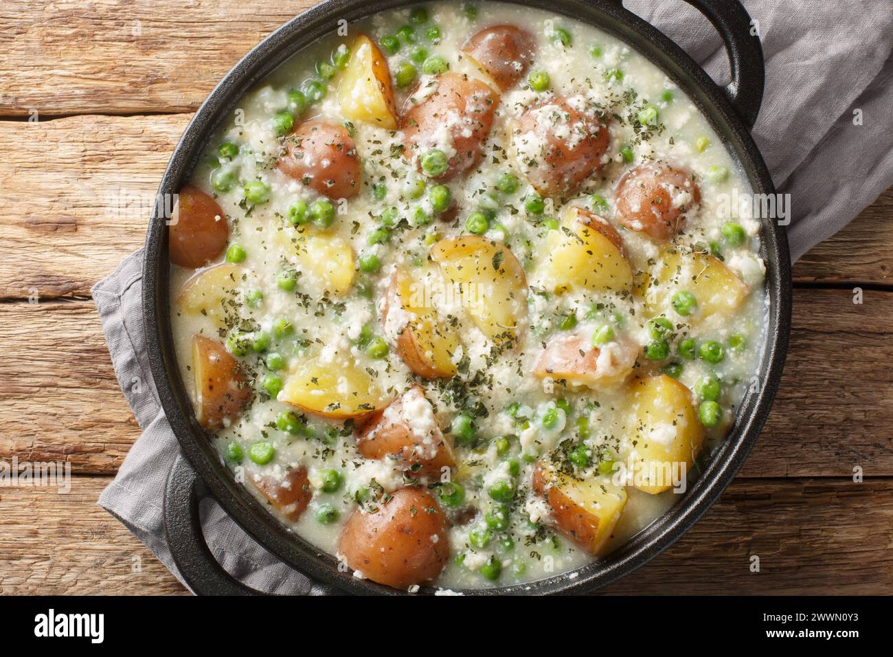 Delicious creamy cheesy potatoes and green peas close-up in a pan on a wooden table. Horizontal top view from above Stock Photo