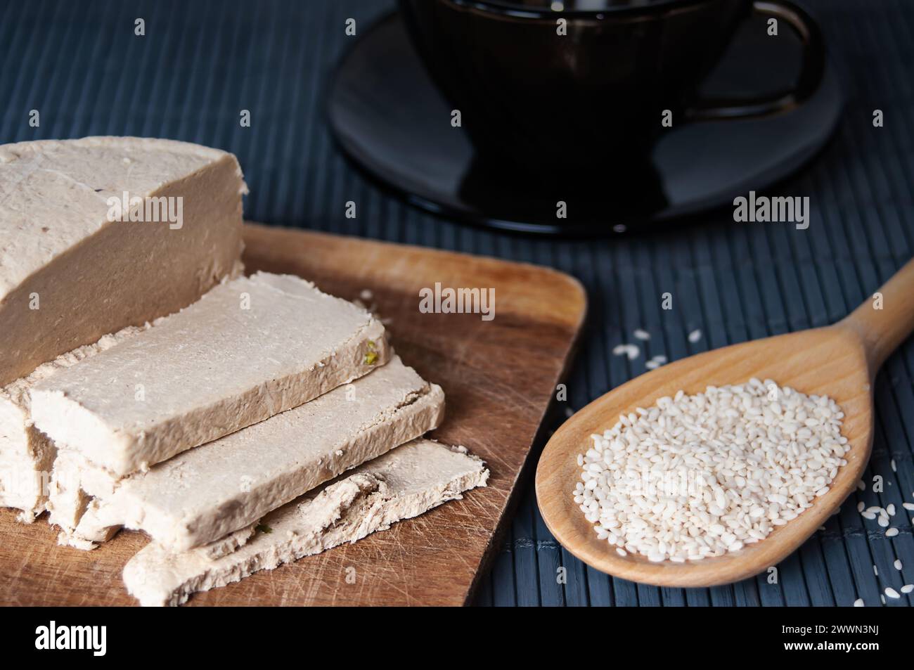 Sliced tahini halva, wooden spoon with sesame seeds and dark glass teacup close up Stock Photo