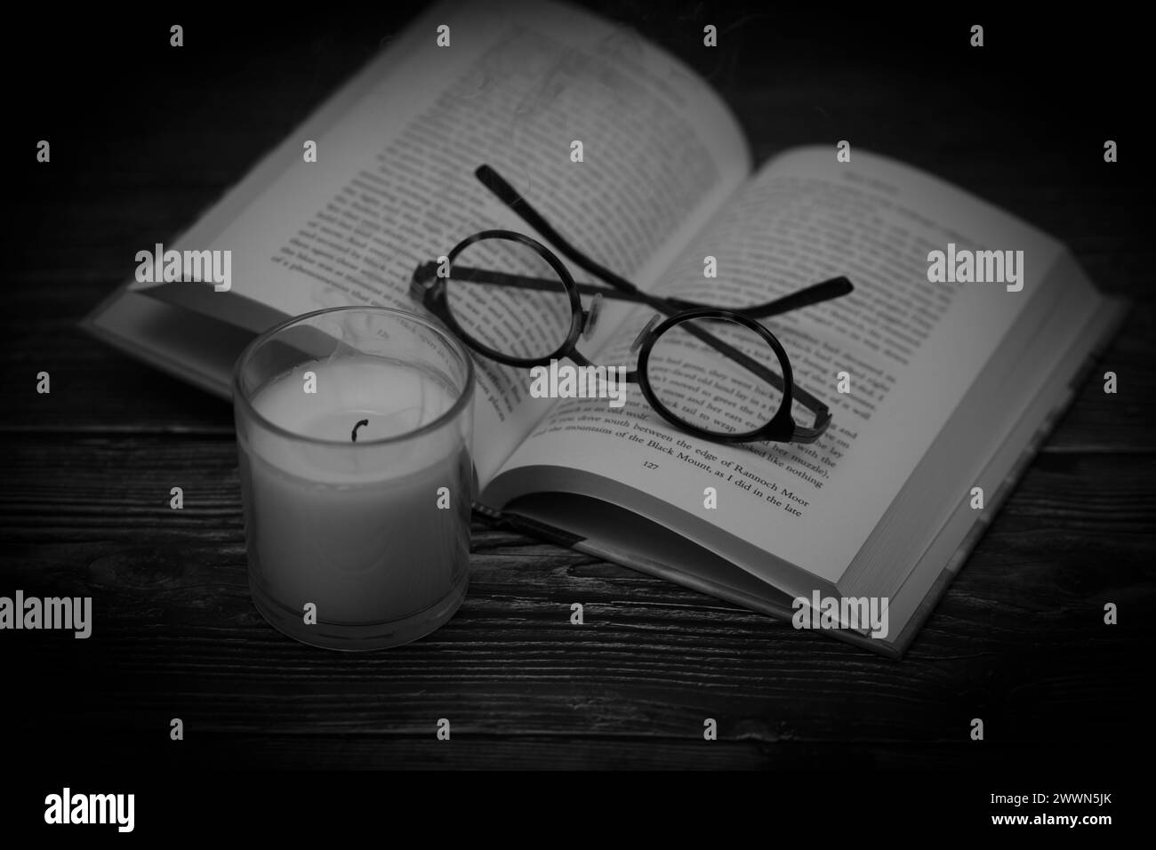 Open hardback book on vintage wooden table with eye glasses folded and recently extinguished candle shot in landscape format, selective focus, mono Stock Photo
