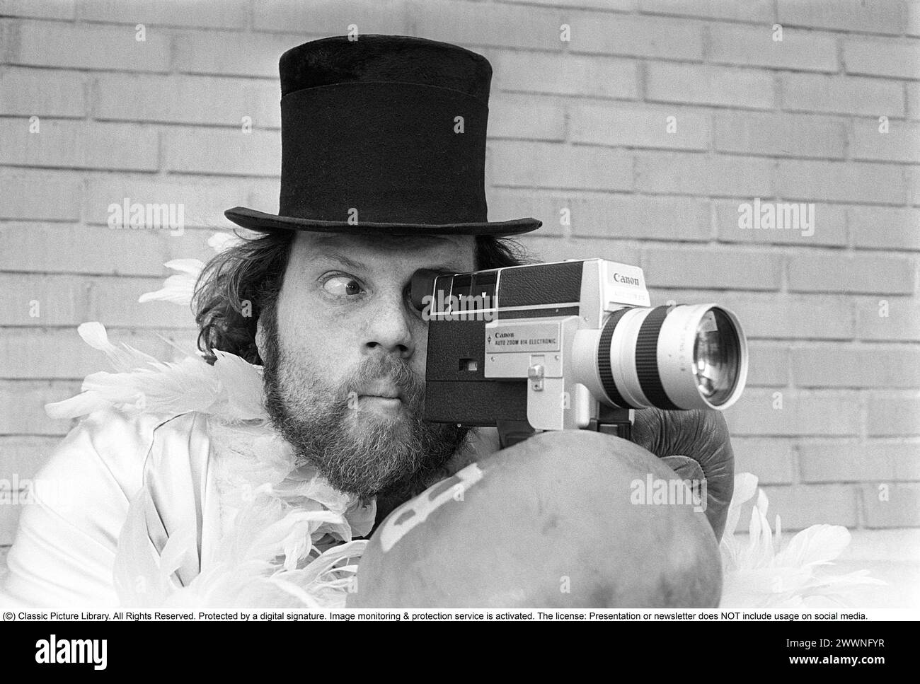 In the 1970s. A man is filming with a amateur film camera. The film was then developed and shown on a film projector on a fold up screen at home. Ricky Bruch (1946-2011) Swedish track and field athlete. He was known for being somewhat of a showman pictured here wearing boxing gloves, a feather boa and a cylinder hat.  1975 Stock Photo