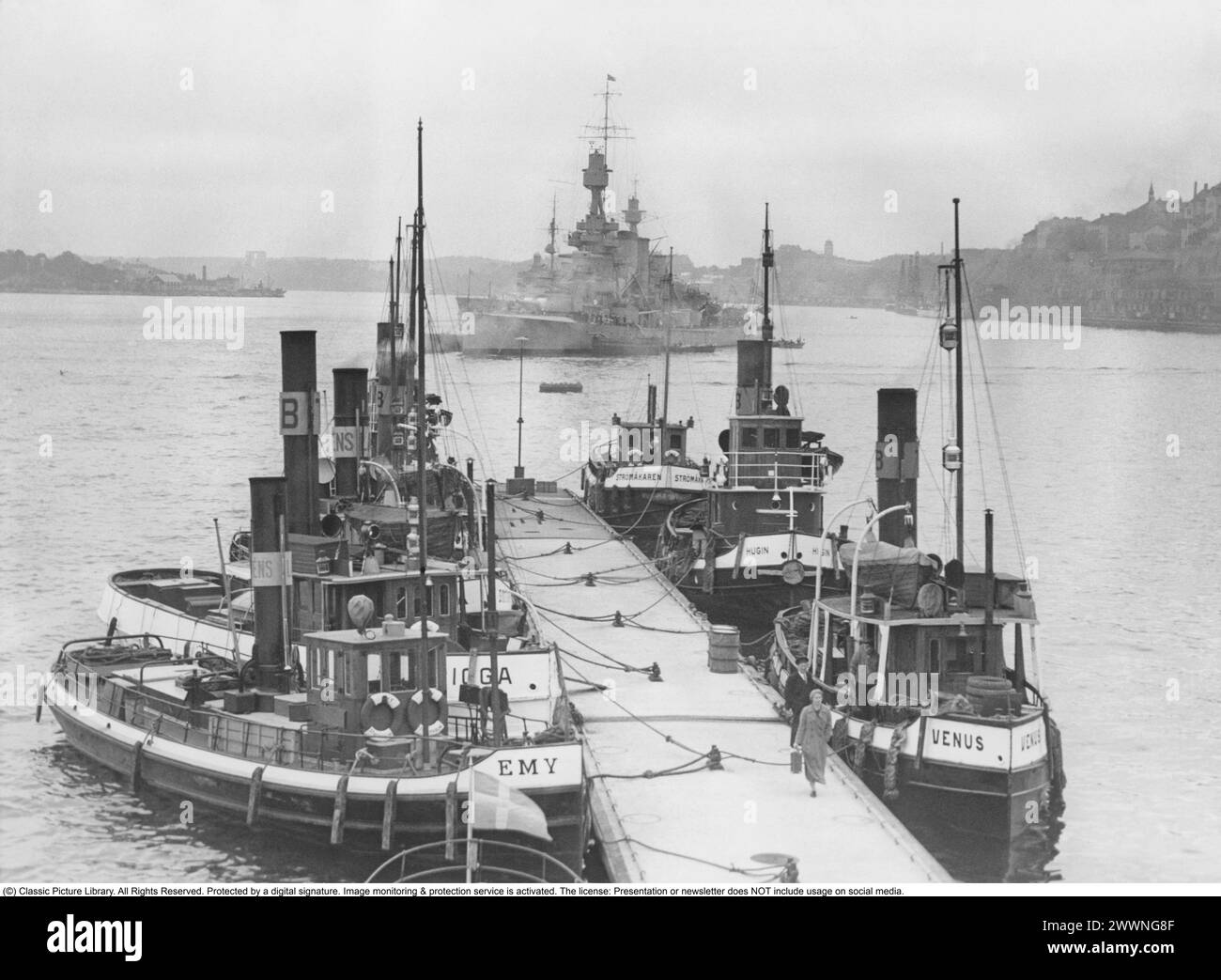 Stockholm harbor in 1937. View of Stockholm harbor and harbor entrance. At Slussen and the jetty, there are tugboats of a smaller type with names such as Emy, Venus, Hugin, Strömåkaren. In the background, a warship from the Swedish coastal fleet is at anchor. To the right Stadsgården. Stock Photo