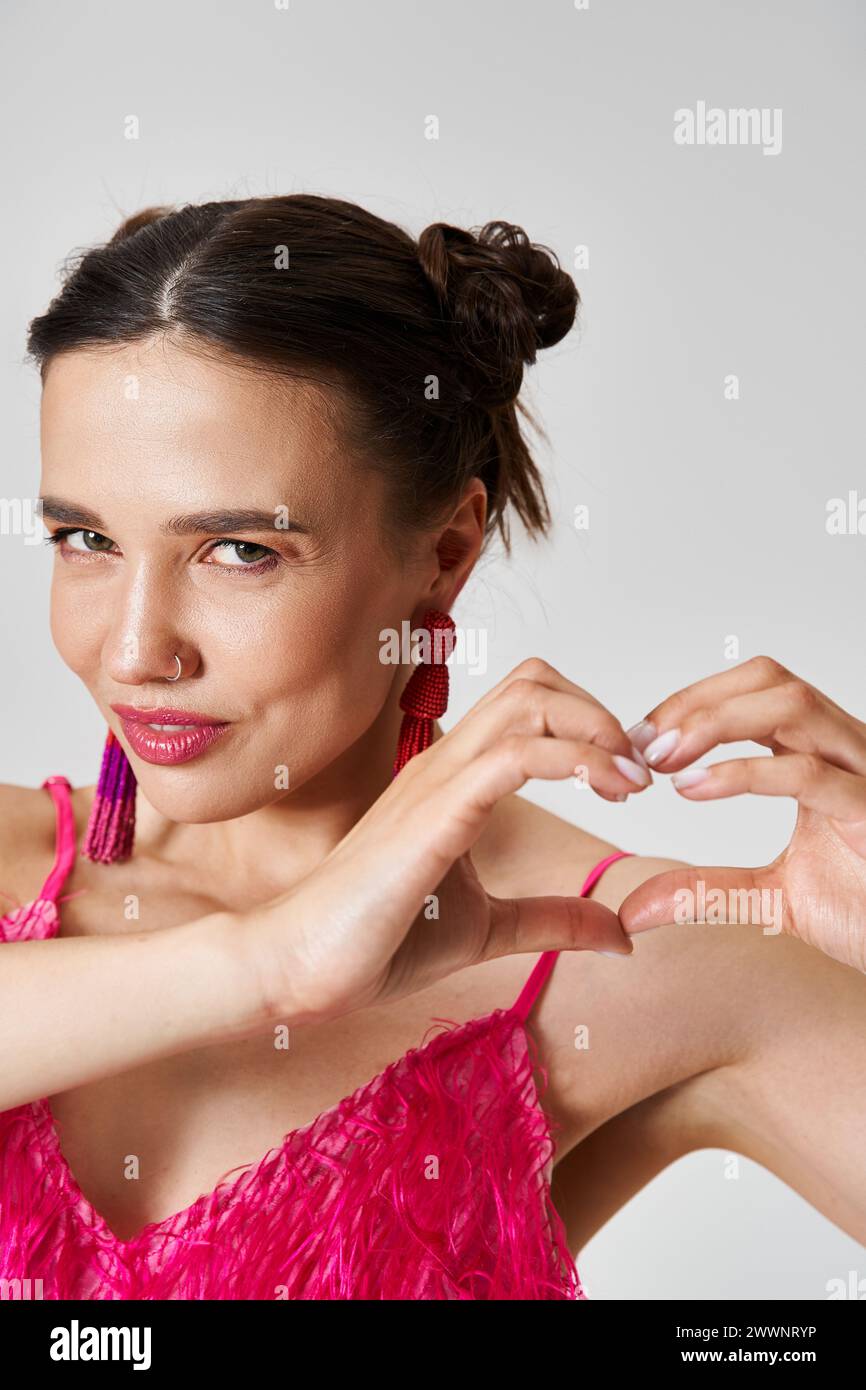 Smiling joyful woman with pierced nose  wearing pink feather dress showing a hand heart to camera Stock Photo