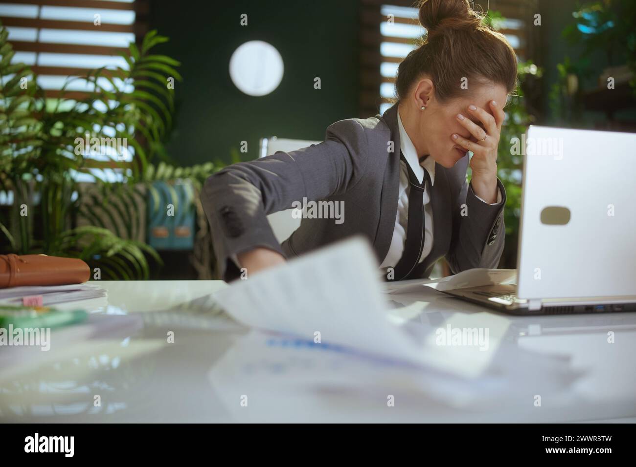 New job. sad modern middle aged woman worker in modern green office in grey business suit with laptop throwing documents. Stock Photo