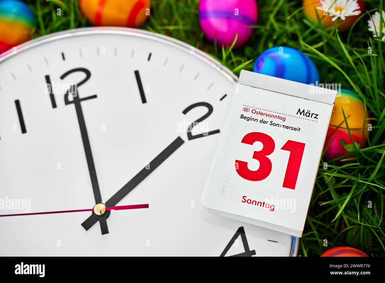 Augsburg, Bavaria, Germany - March 25, 2024: Symbolic image of the time change from winter time to summer time at Easter. A clock lies in a meadow together with colorful Easter eggs and a calendar with the date 31 March, Easter Sunday *** Symbolbild Zeitumstellung von Winterzeit auf Sommerzeit an Ostern. Eine Uhr liegt in einer Wiese zusammen mit bunten Ostereiern und einem Kalender mit Datum 31 März, Ostersonntag Stock Photo