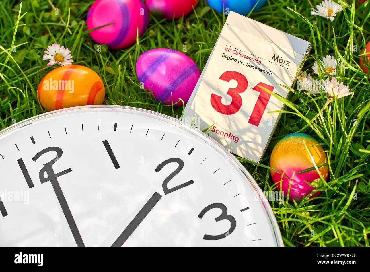 Augsburg, Bavaria, Germany - March 25, 2024: Symbolic image of the time change from winter time to summer time at Easter. A clock lies in a meadow together with colorful Easter eggs and a calendar with the date 31 March, Easter Sunday *** Symbolbild Zeitumstellung von Winterzeit auf Sommerzeit an Ostern. Eine Uhr liegt in einer Wiese zusammen mit bunten Ostereiern und einem Kalender mit Datum 31 März, Ostersonntag Stock Photo