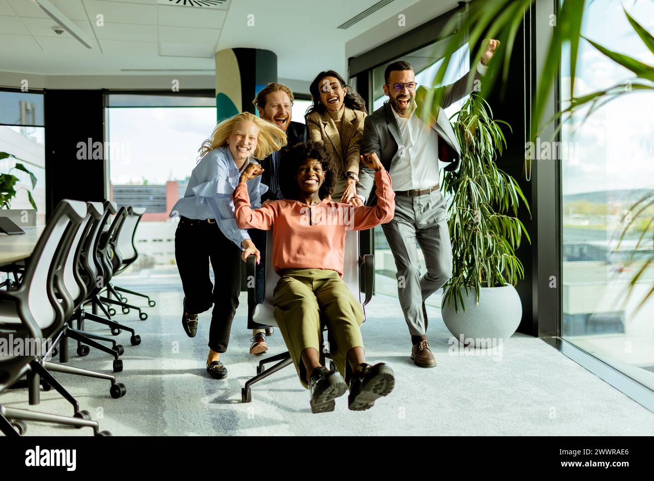 Team of coworkers shares a moment of fun, racing an office chair amidst a well-lit workspace. Stock Photo