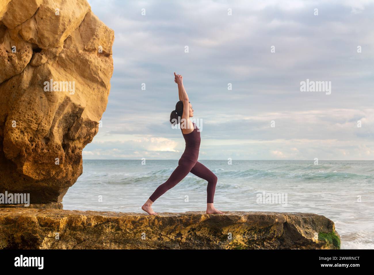 Woman practicing yoga standing on a rock by the ocean, arms above head stretching. Stock Photo