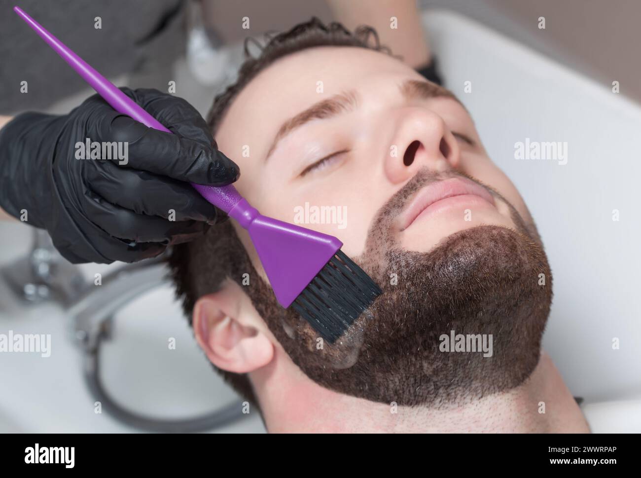 The barber paints the beard and the mustache of the young man in the barbershop. Stock Photo