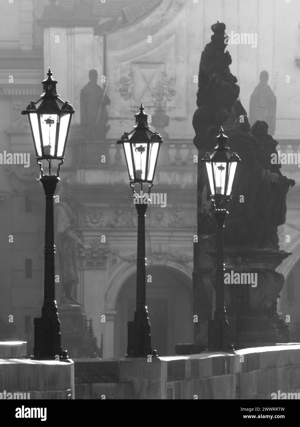 Street lamps on Charles bridge illuminated by morning sun and dark silhouettes of statues, Prague, Czech Republic. Black and white image. Stock Photo