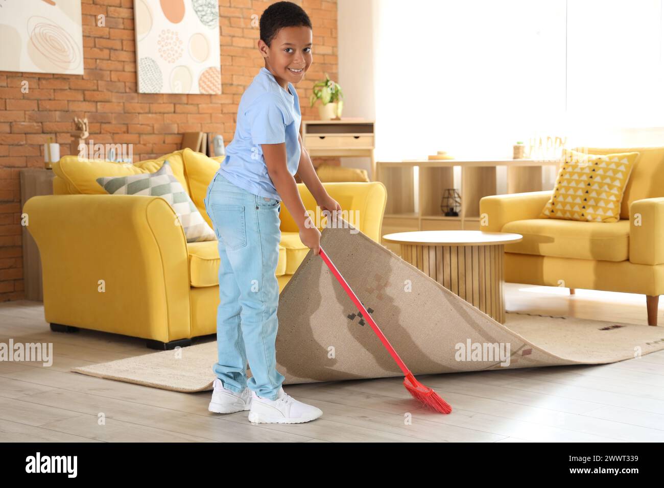 Cute AfricanAmerican boy sweeping dust under carpet with broom in