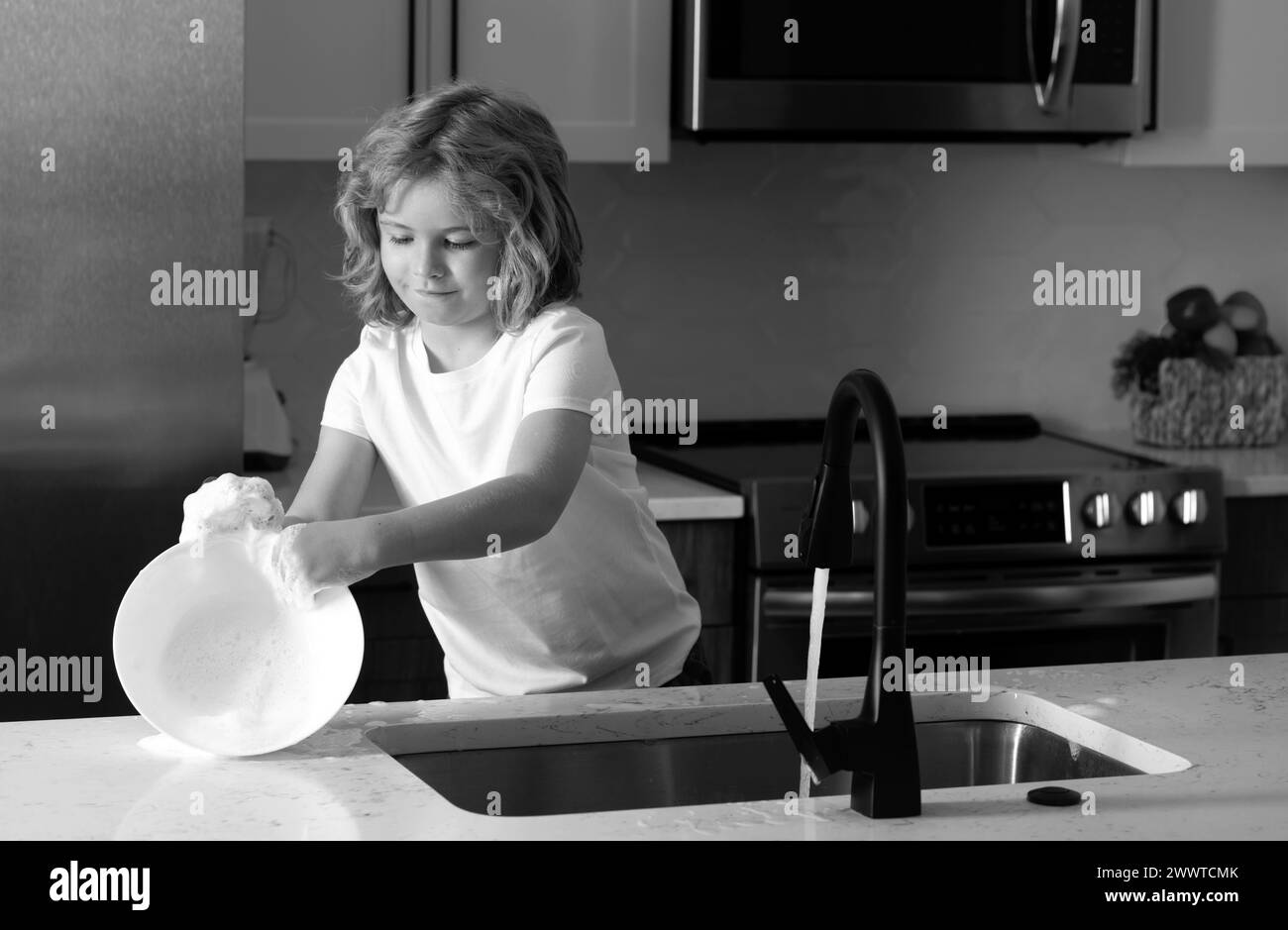 Child housekeeper washing the dishes on soapy water. Cute Funny boy washing dishes in kitchen. American kid learning domestic chores at home. Cleaning Stock Photo