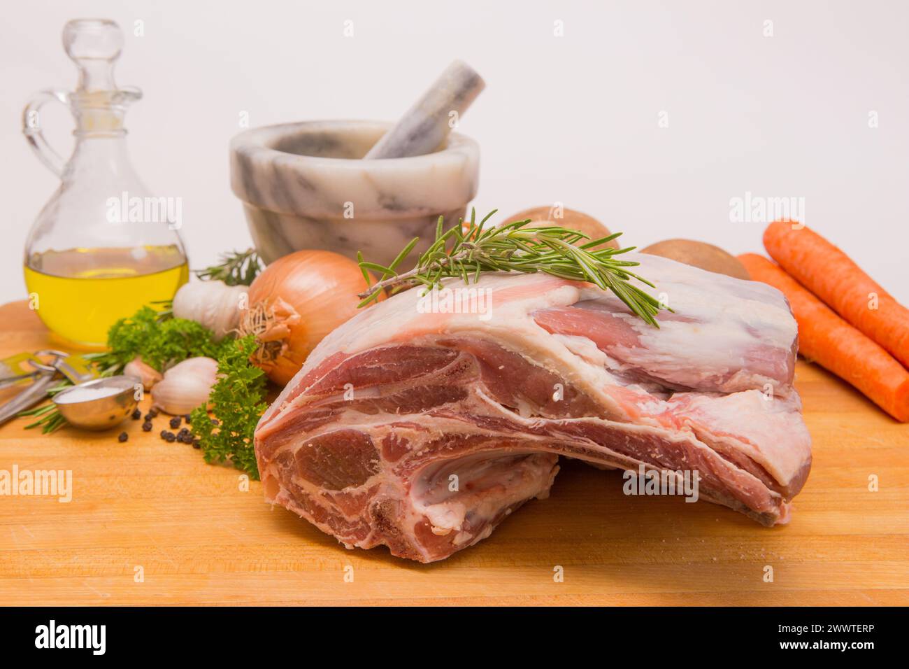 Fresh raw Shoulder of lamb on a wood cutting board with fresh herbs, salt and peppercorns , olive oil, onions, potatoes and a mortar and pestle Stock Photo
