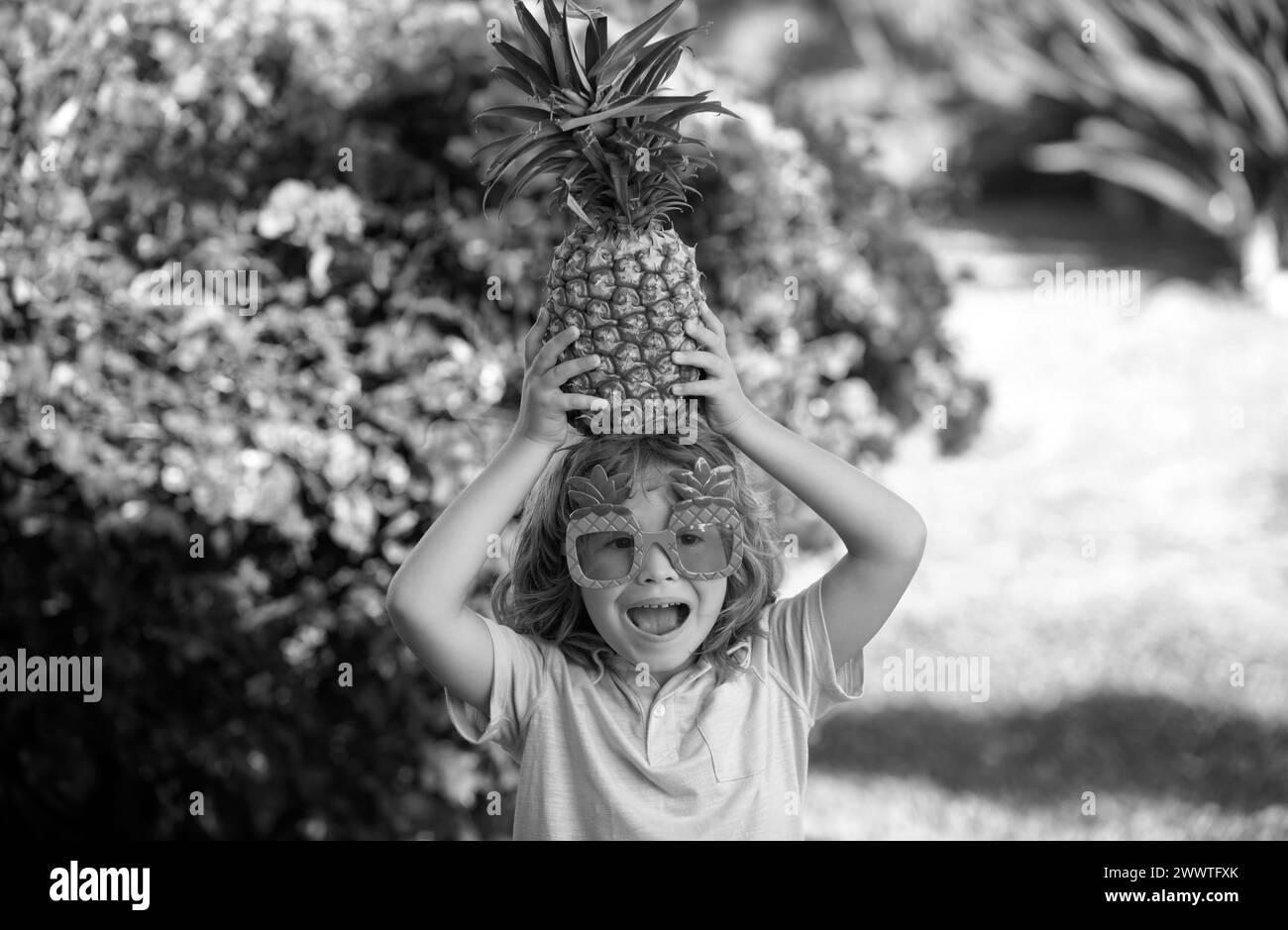 Funny child holds a pineapple on her head on a summer nature background. Pineapple kid boy and pineapples on summer vacation. Stock Photo