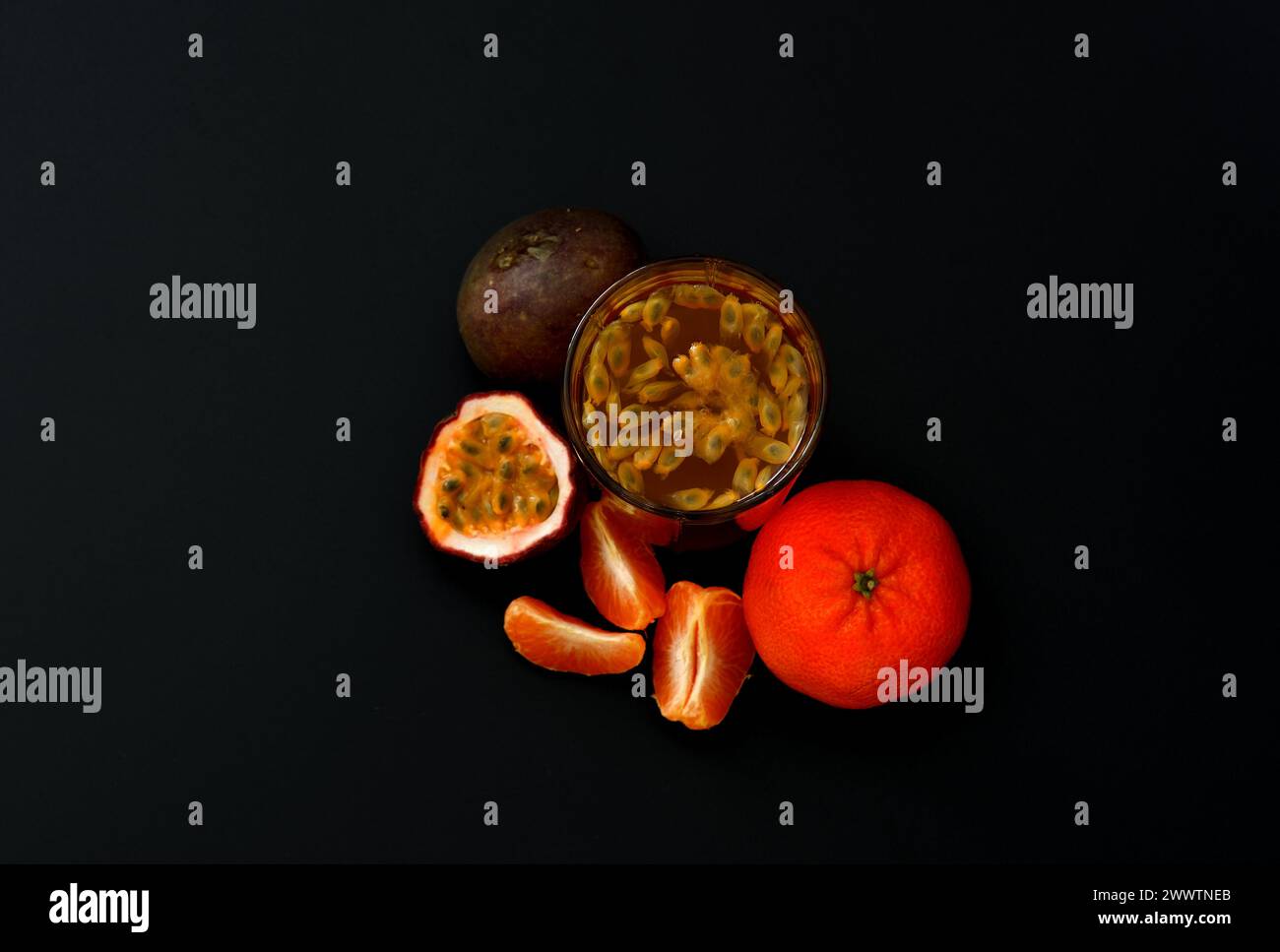 A glass of fruit juice with seeds on a black background, next to pieces of ripe mandarin passion fruit. Top view, flat lay. Stock Photo