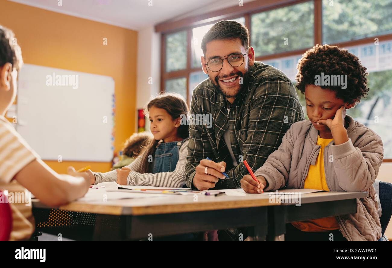 Primary educator smiling at the camera in a classroom. Teacher teaching a group of children in a school. Man giving an art lesson in an early child de Stock Photo
