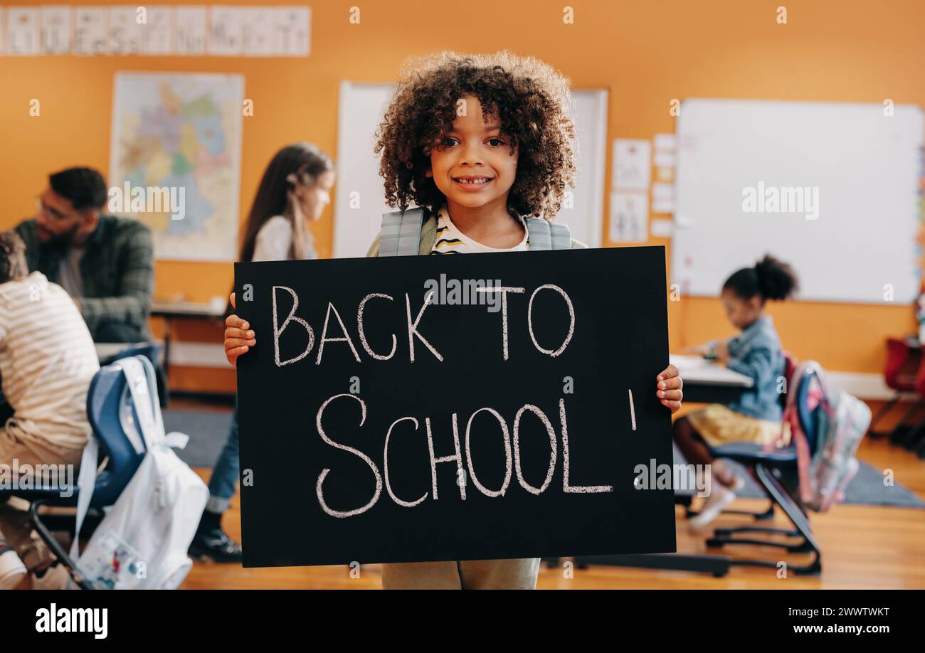 Proud pupil standing in front of co-ed classroom on first day of elementary school, holding a back to school sign. Boy excited to start a new class wi Stock Photo
