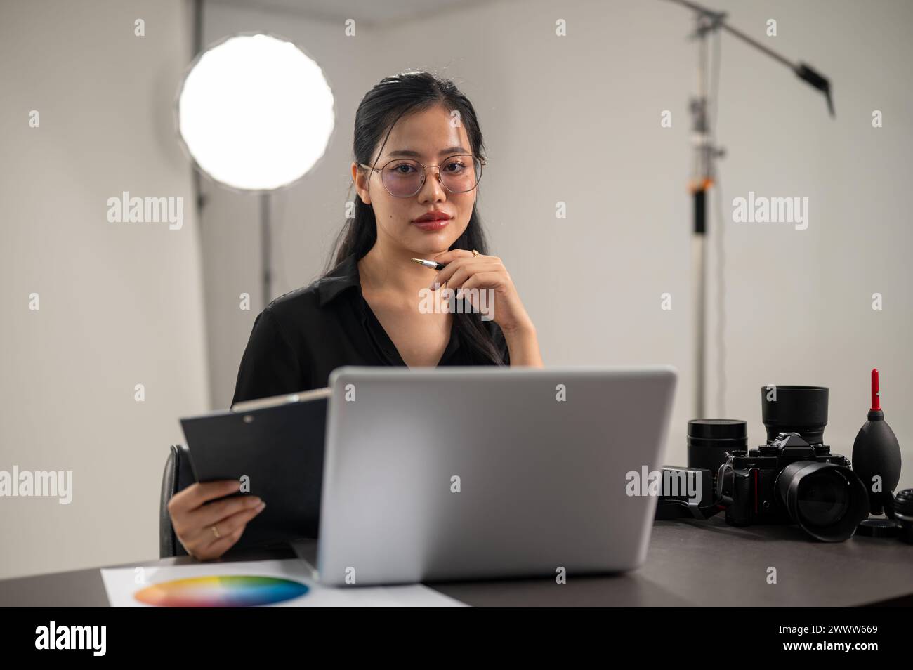 An attractive, confident Asian female photographer is looking at the camera while sitting at a desk in a photoshoot studio. Stock Photo