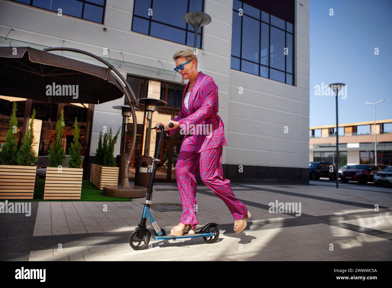 Stylishly dressed mature woman in a bright pink pantsuit of trousers and a jacket rides around the city on a scooter, an active, healthy and eco frien Stock Photo