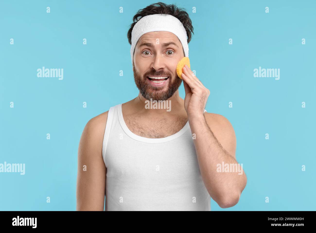Man with headband washing his face using sponge on light blue background Stock Photo