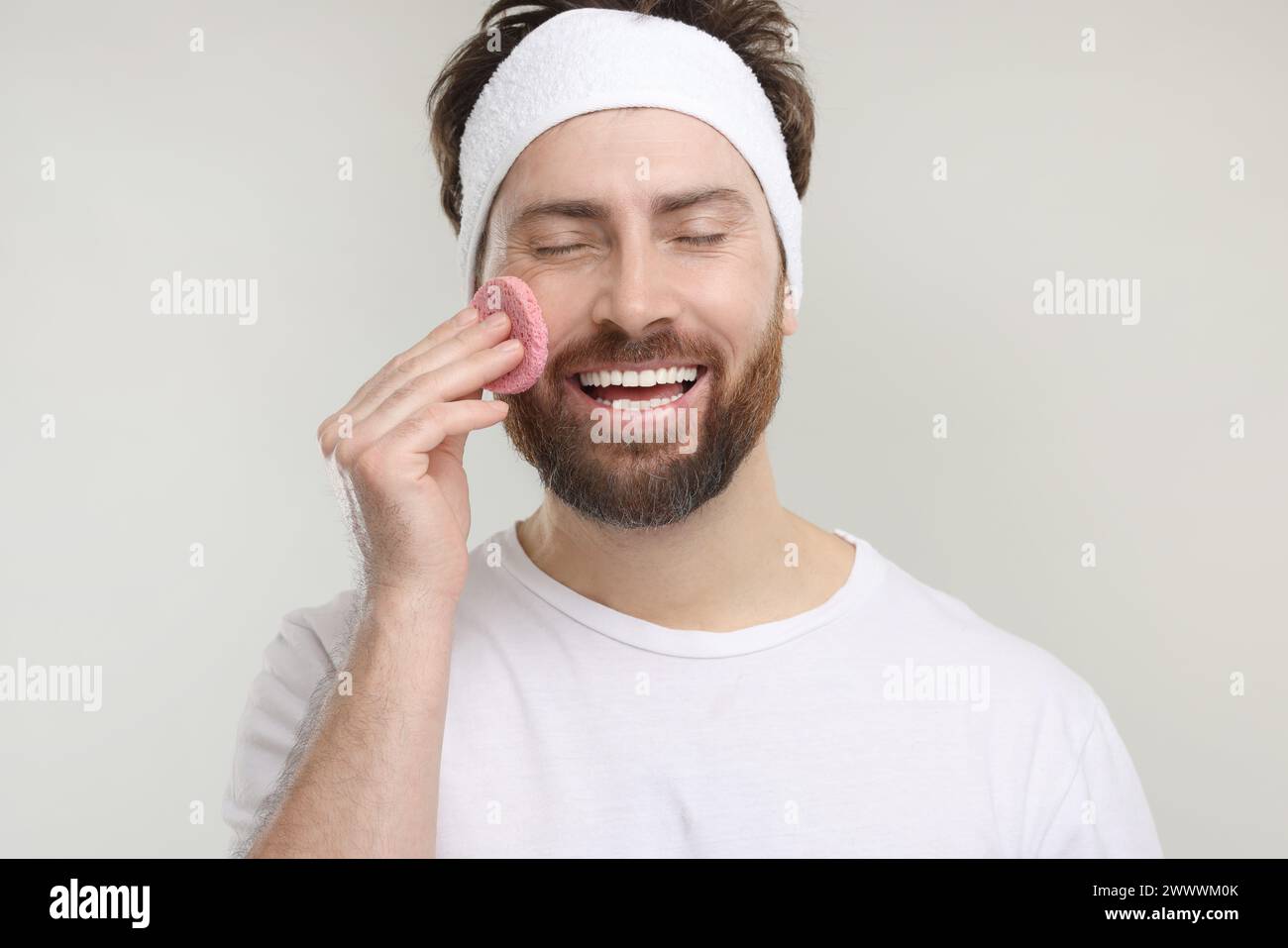 Man with headband washing his face using sponge on light grey background Stock Photo