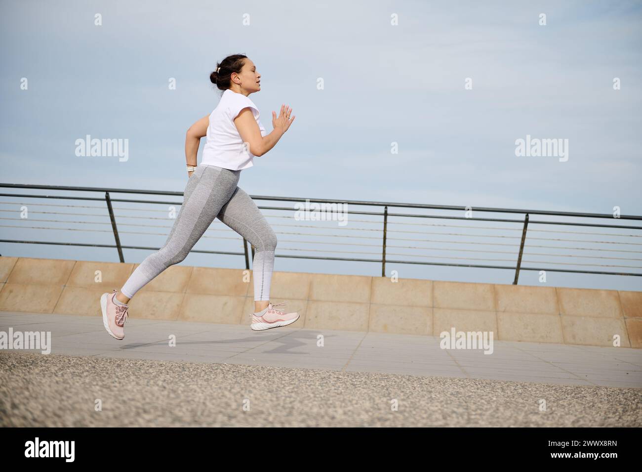 Side portrait of woman running on the city bridge, performing her morning jog, exercising outdoors. Athletic female burning calories nd loosing weight Stock Photo