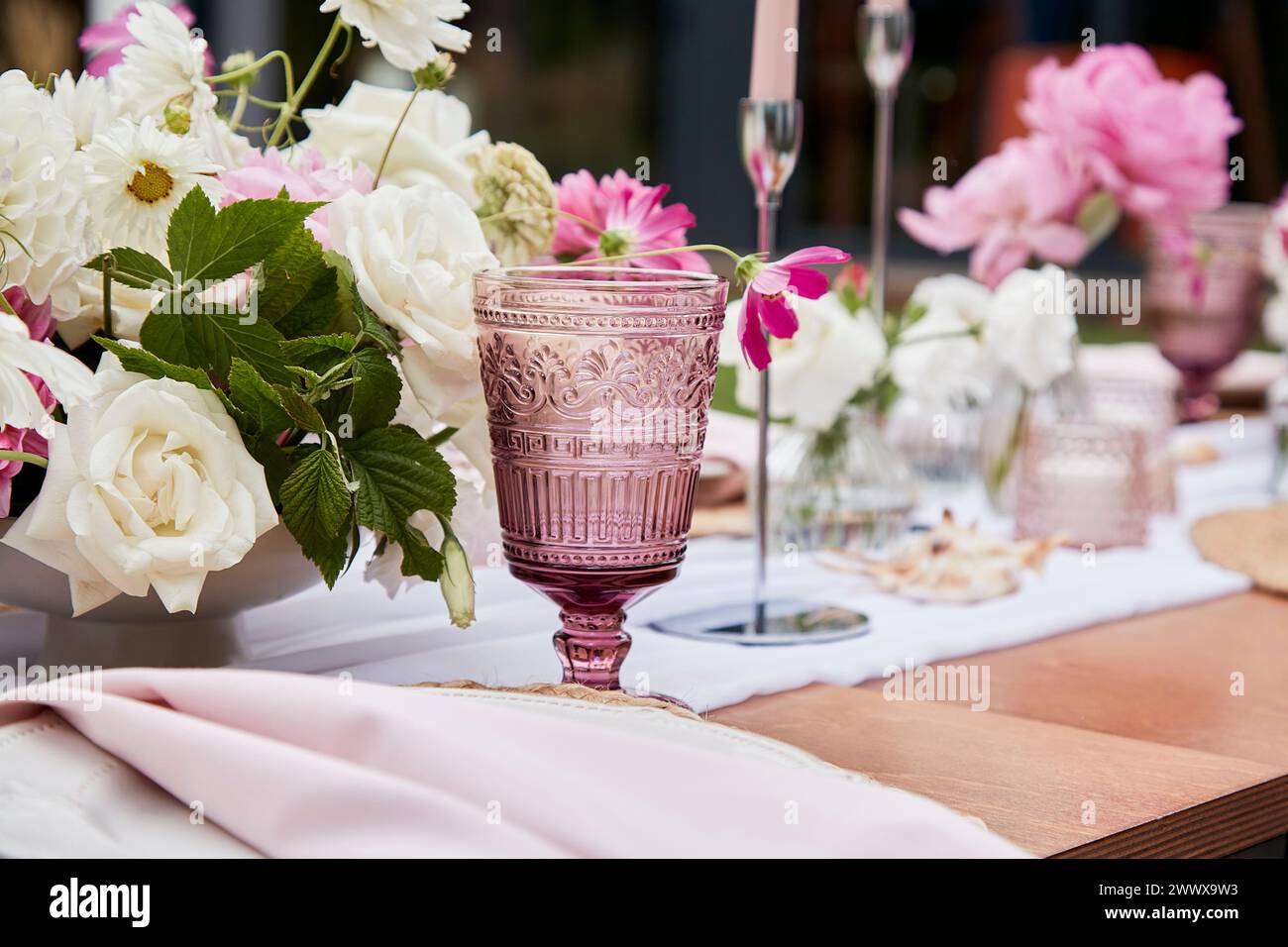 Outside aesthetic festive table setting among pink flowers arrangement, candles and shells decor. Stock Photo